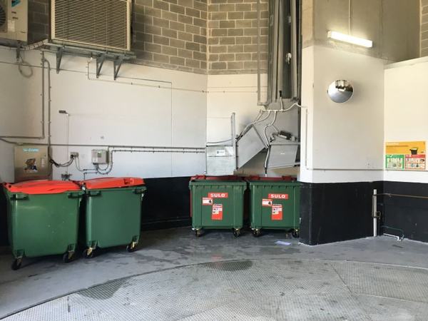 A bin store with green bins along the wall, a Garbage Doctor unit and its conduit running above them to the chute