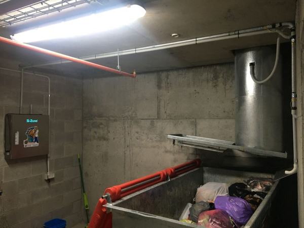 A garbage chute discharging into a steel bin full of rubbish bags, with a Garbage Doctor unit on the wall and its pipe into the bin