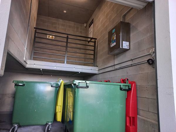 Green bins with yellow and red lids in a bin room, under a Garbage Doctor mounted high on the wall