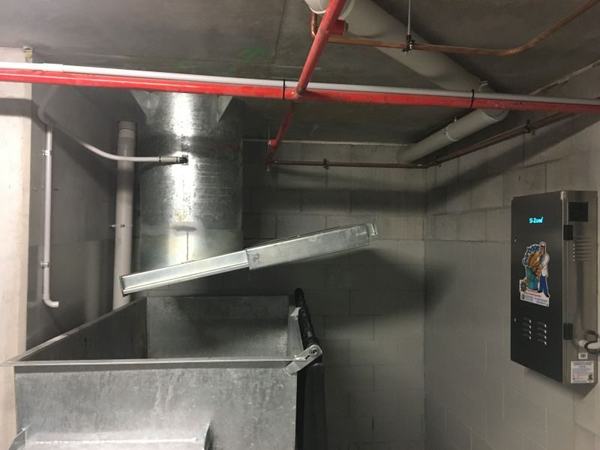 A large steel bin under a garbage chute's discharge, with a Garbage Doctor on the wall beside it
