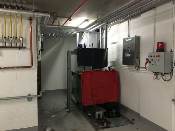 The bin lifter and its warning light in a compactor room, with a Garbage Doctor on the wall beside them