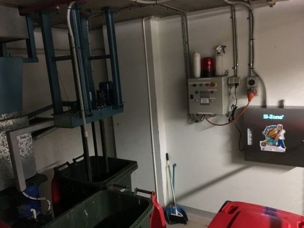 A compactor room with a Garbage Doctor on its wall, next to the compactor's control panel and bin lifter