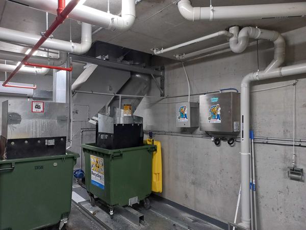 A Garbage Doctor Duo in a basement bin room, with grey conduit along the wall above the bins and the chute's hopper beside them