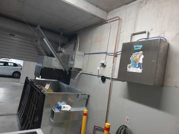 A steel bin under a garbage chute's hopper, with a Garbage Doctor Duo, its conduit and its air hose on the wall beside it