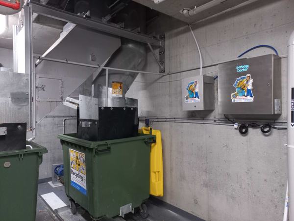 A basement bin room with a Garbage Doctor Duo on the wall, its conduit leading to the bin under the garbage chute