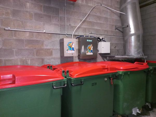 A row of red-lidded bins under a Garbage Doctor Duo and its control box, on a block wall beside the chute