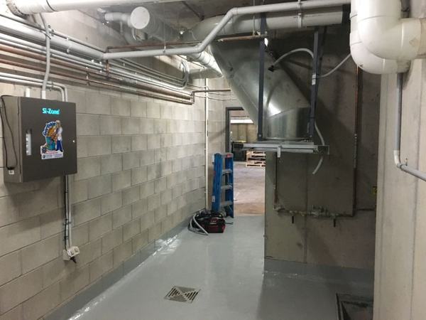 A basement service corridor with a Garbage Doctor beside the chute's discharge, and its conduit along the wall