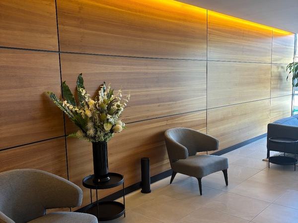 A slim black Scent Doctor tower on the floor of a lobby, beside a vase of flowers and two armchairs, against a timber-panelled wall
